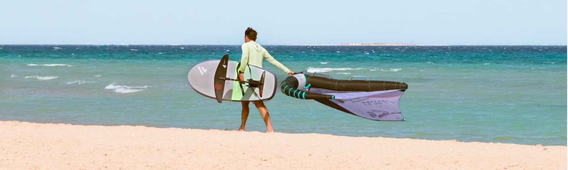 Surfer trägt Brett und Segel am Strand am ION Club Safaga entlang.