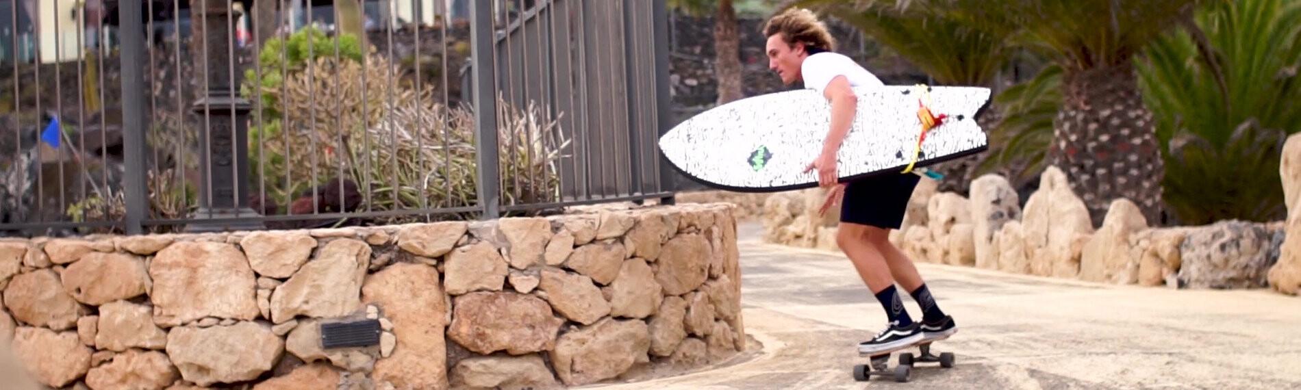 Person auf Longboard mit Surfbrett unter dem Arm, fährt nahe eines gemauerten Steingeländers bei Palmen.
