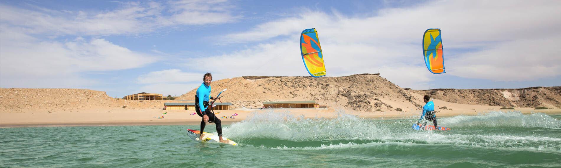 Kitesurfer auf dem Wasser mit farbigen Schirmen vor einer Wüstenkulisse in Dakhla, Marokko