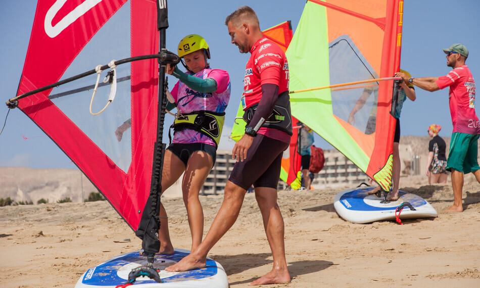 Menschen beim Windsurf-Training am Sandstrand, mit bunten Segeln und Boards, tragen Neoprenanzüge und Helme vor dem René Egli Center auf Fuerteventura.