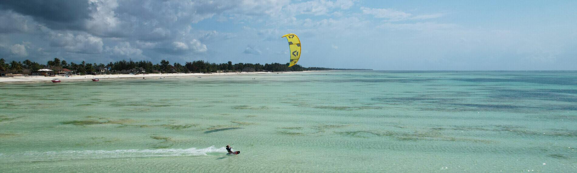 Ein Kitesurfer gleitet über das türkisfarbene Meer, während ein gelber Kite vor einem bewölkten Himmel sichtbar ist.