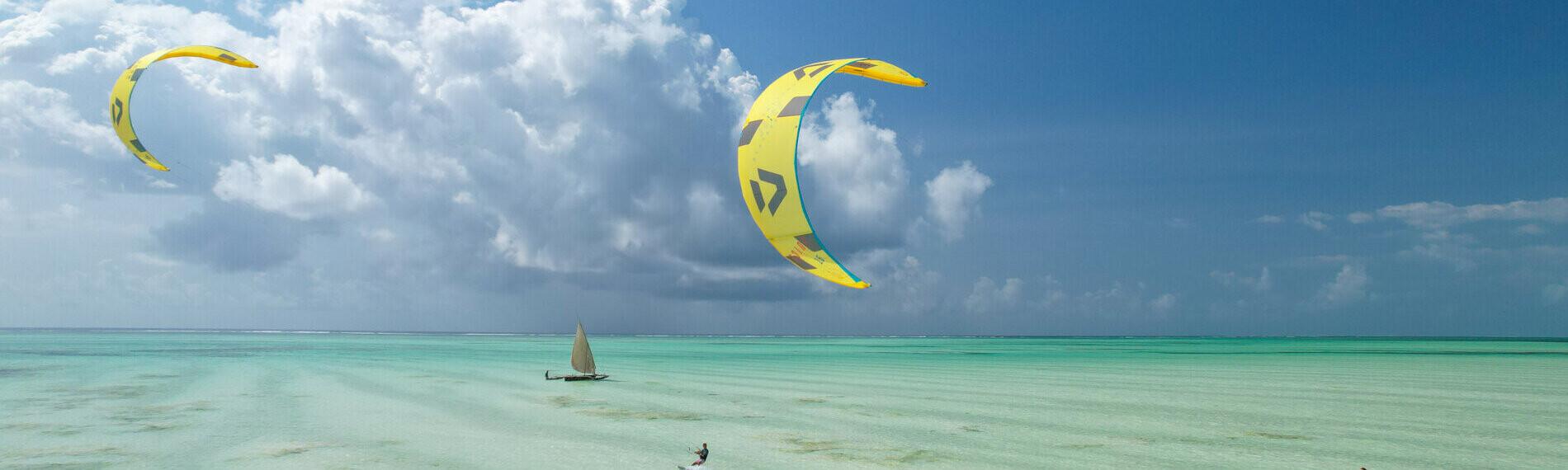 Kitesurfer auf kristallklarem Wasser unter blauem Himmel mit Wolken, zwei gelbe Kites segeln im Wind.