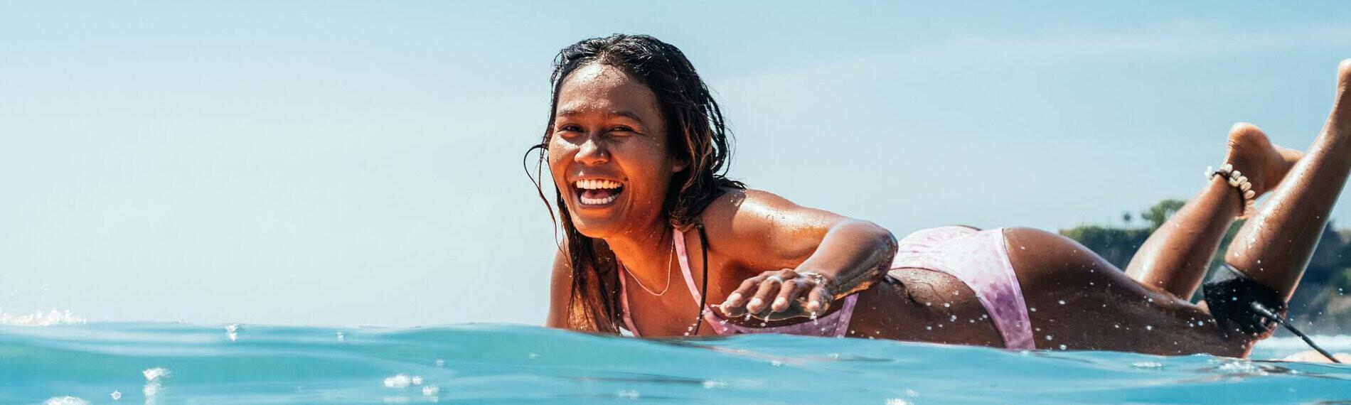 Frau auf Surfbrett lächelnd im Meer liegend, sonniger Himmel im Hintergrund.