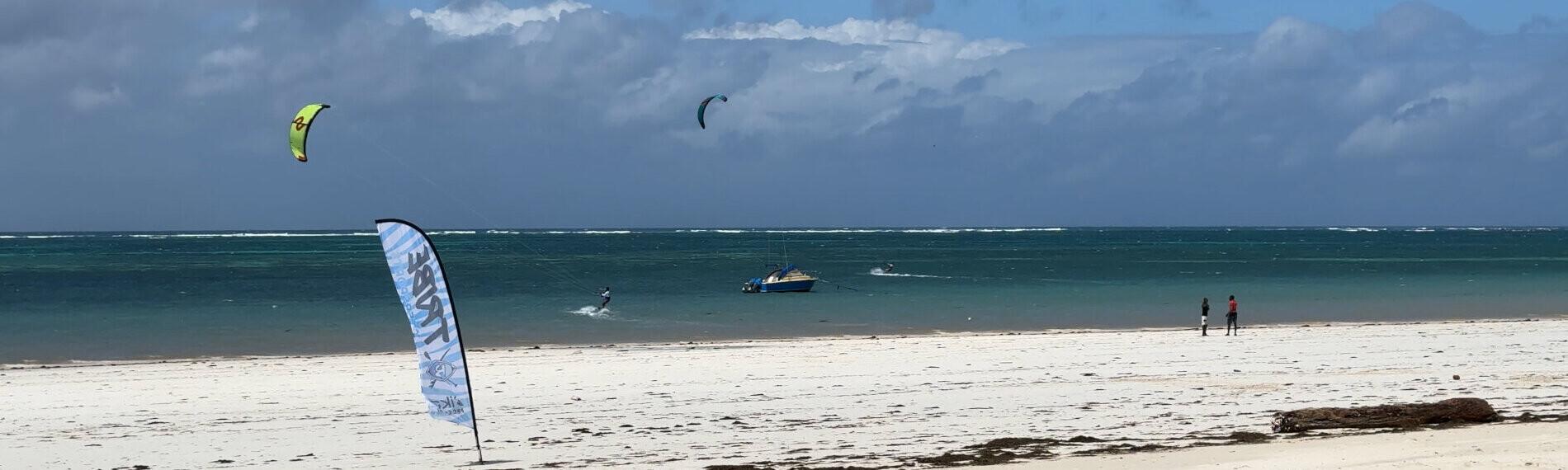 Kenia Strand mit Kite-Surfern im Hintergrund, blauem Himmel, wenigen Wolken und einer kleinen Fahne im Vordergrund.
