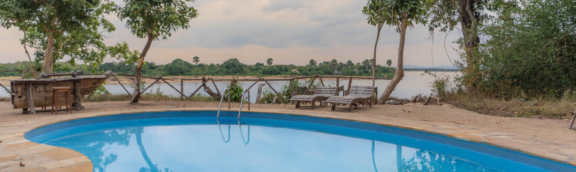 Tansania Landsafari - Selous River Camp Runder Swimmingpool mit Steinterrasse, umgeben von Bäumen. Im Hintergrund ist ein Fluss und bewölkter Himmel zu sehen.