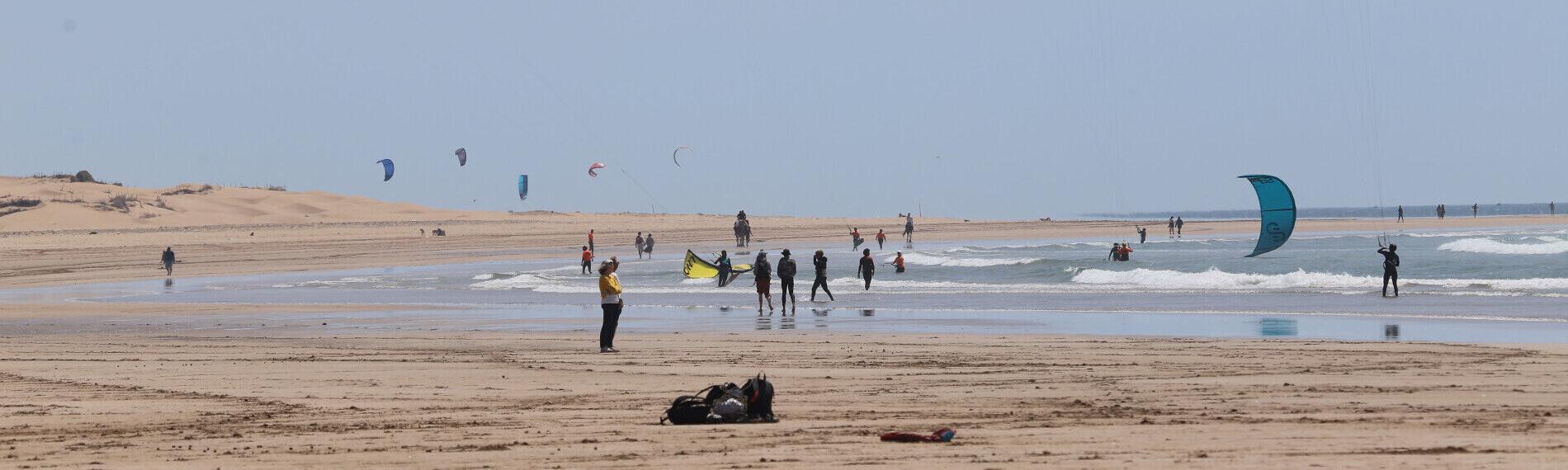 Essaouira - Salt Water Elements - Kitesurfer am Strand mit bunten Kites bei leicht bewölktem Himmel und sandiger Küstenlinie im Hintergrund.